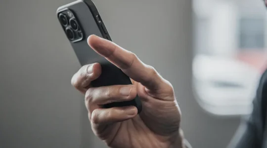 Main tenant un smartphone en position naturelle, le pouce cherchant a atteindre des elements situes trop haut sur l ecran, avec un fond flou et de l espace negatif pour un titre editorial.